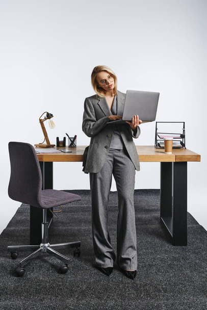 привлекательная элегантная деловая женщина в сером костюме позирует рядом со столом с ноутбуком и смотрит в камеру - Фото, изображение