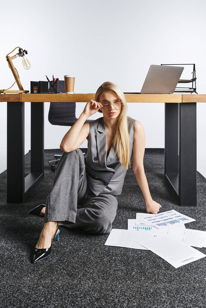 debonair blonde woman in elegant gray suit working on floor with her papers and looking at camera - Фото, изображение