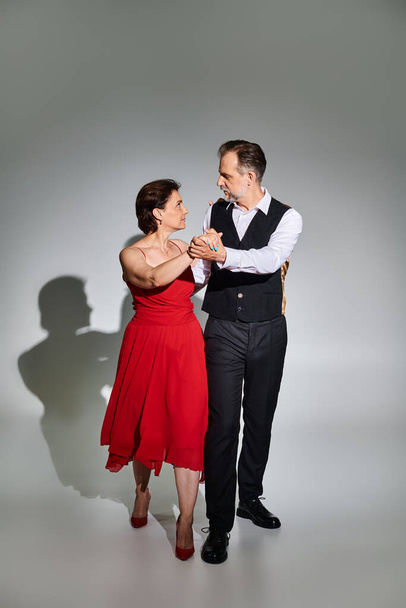 Повнометражне зображення зрілої привабливої пари в червоній сукні та костюмі, що танцює на сірому фоні - Фото, зображення