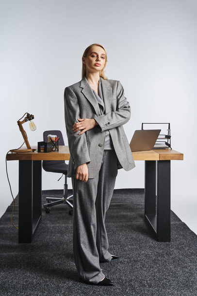 atrakcyjna ciężko pracująca bizneswoman w wyrafinowanym szarym garniturze patrząc na kamerę na szarym tle - Zdjęcie, obraz
