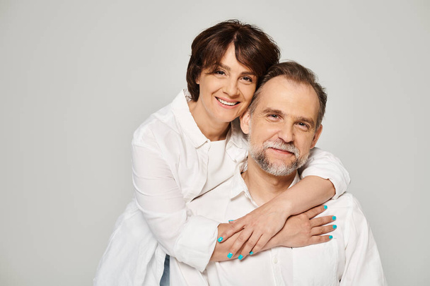 Retrato de pareja sonriente de mediana edad abrazando y mirando a la cámara sobre fondo gris - Foto, Imagen