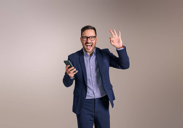 Mladý podnikatel s chytrým telefonem nadšeně křičí a gestikuluje OK znamení na bílém pozadí - Fotografie, Obrázek