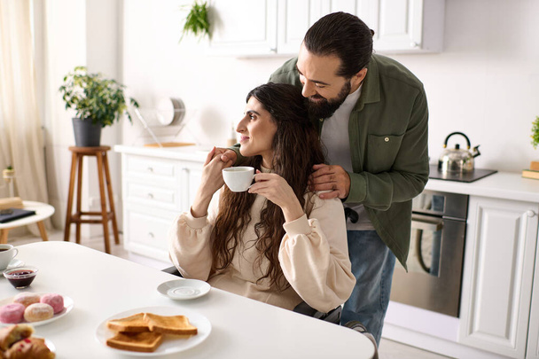 bärtiger liebevoller Ehemann umarmt seine behinderte hübsche Frau beim Frühstück in der heimischen Küche - Foto, Bild