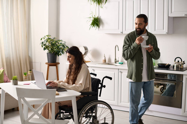 handsome man drinking coffee next to his jolly disabled wife in wheelchair working at laptop - Фото, зображення