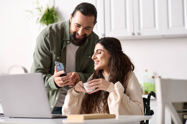appealing loving couple of bearded man and disabled woman looking at phone in kitchen at home - Photo, Image