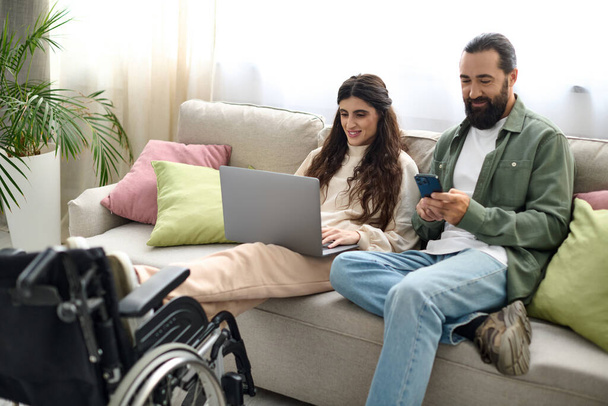 hermosa mujer con discapacidad de movilidad sentado en el sofá con el ordenador portátil junto a su marido con teléfono - Foto, imagen