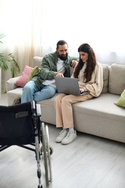 piękna kobieta z niepełnosprawnością ruchową siedzi na kanapie z laptopem obok męża z telefonem - Zdjęcie, obraz