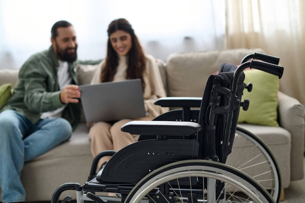 zaměřit se na moderní invalidní vozík před rozmazaným vousatým mužem a postiženou ženou dívající se na notebook - Fotografie, Obrázek
