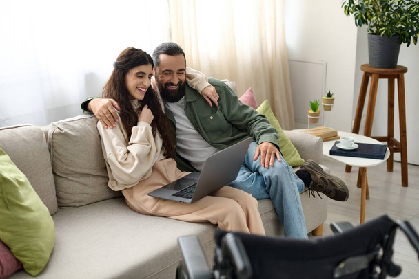 loving couple of bearded man and disabled beautiful wife spending time together at laptop on sofa - Photo, Image