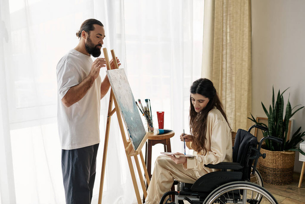 όμορφος γενειοφόρος άνδρας βλέποντας τα άτομα με ειδικές ανάγκες όμορφη γυναίκα του σε αναπηρική καρέκλα ζωγραφική στο καβαλέτο στο σπίτι - Φωτογραφία, εικόνα