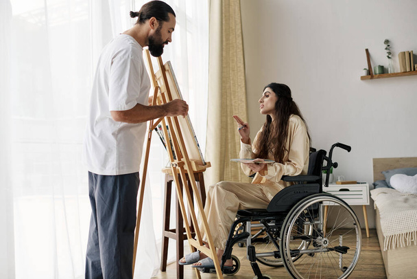 schöner bärtiger Mann beobachtet seine behinderte schöne Frau im Rollstuhl beim Bemalen auf der Staffelei zu Hause - Foto, Bild