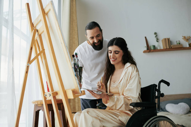 ελκυστικός γενειοφόρος βλέποντας τα άτομα με ειδικές ανάγκες όμορφη γυναίκα του σε αναπηρική καρέκλα ζωγραφική στο καβαλέτο στο σπίτι - Φωτογραφία, εικόνα
