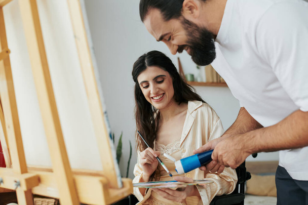 люблячий веселий чоловік допомагає своїй інклюзивній хорошій дружині малювати на мольберті вдома - Фото, зображення