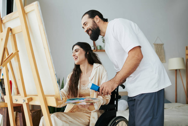 любящий веселый муж помогает своей инклюзивной красивой жене рисовать на мольберте, находясь дома - Фото, изображение
