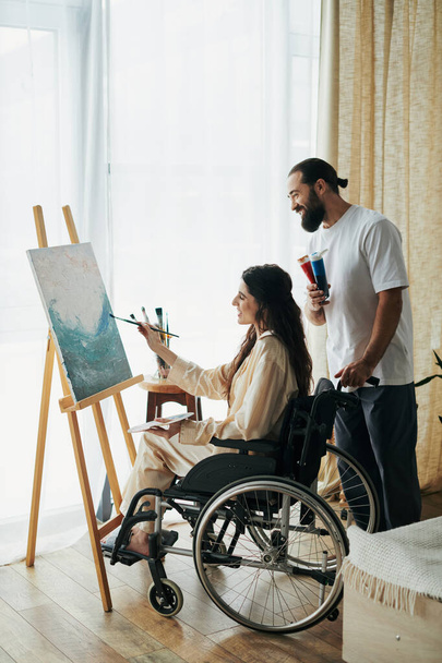 good looking bearded man watching disabled beautiful wife on wheelchair painting on easel at home - Photo, Image
