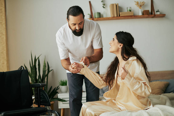 весёлый любящий мужчина в домашней одежде помогает своей жене-инвалиду переодеться в пижаму в спальне дома - Фото, изображение
