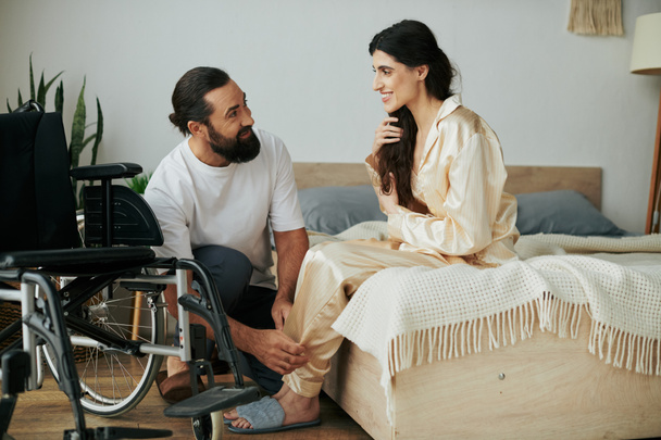 бородатый любящий мужчина в домашней одежде помогает своей жене-инвалиду переодеться в пижаму в спальне дома - Фото, изображение