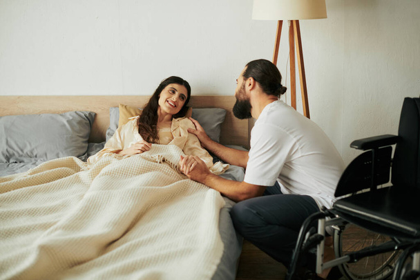 piękna radosna kobieta z niepełnosprawnością ruchową leżąca w łóżku obok swojego brodatego kochającego męża - Zdjęcie, obraz