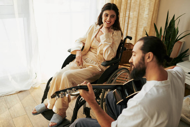 donna allegra con disabilità in sedia a rotelle a guardare il marito barbuto suonare la chitarra per lei - Foto, immagini