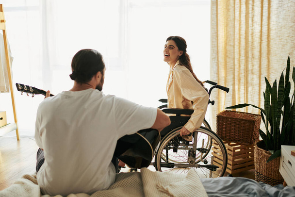 весёлая женщина с инвалидностью в инвалидном кресле наблюдает, как её бородатый муж играет для неё на гитаре. - Фото, изображение