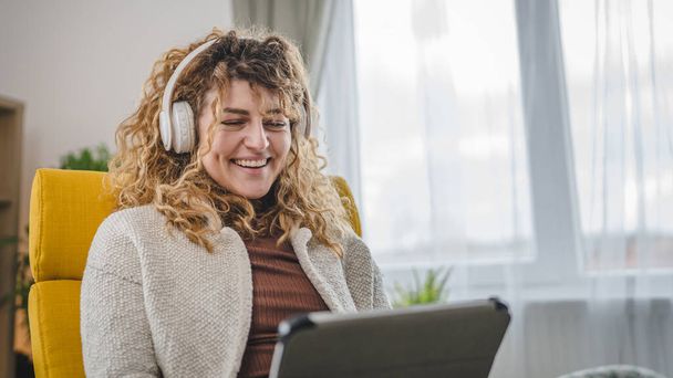 Jedna kaukaska kobieta z kręconymi włosami siedzieć w domu za pomocą tabletu cyfrowego i słuchawek do oglądania filmów lub seriali online strumienia lub mieć połączenie wideo szczęśliwy uśmiech mieć dobry czas przestrzeń kopiowania - Zdjęcie, obraz