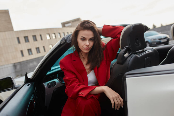 Elegante joven morena en traje de pantalón rojo, sentado en el asiento del conductor con la puerta abierta del coche convertible blanco en el aparcamiento al aire libre, mirando con confianza a la cámara - Foto, imagen