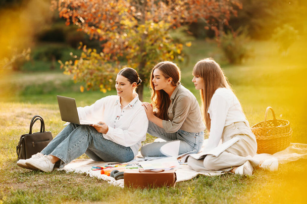 Tres mujeres jóvenes que aprenden juntas en línea desde una computadora portátil mientras están sentadas en el parque sobre una manta. - Foto, Imagen