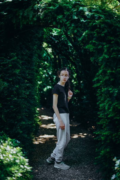 Fransa 'da güneşli bir yaz gününde botanik bahçesinde çiçek kemeriyle yol kenarında dikilen gözlüklü ve spor giysili güzel beyaz bir genç kızın portresi.. - Fotoğraf, Görsel