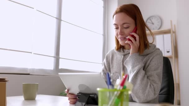 Nuori nainen puhuu matkapuhelimella käyttäessään digitaalista tablettia, joka istuu työpöydällä kotitoimistossa. Pienyritysten ja teknologian elintapojen käsite. - Materiaali, video