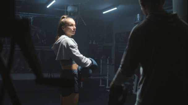 Athletic fighter in boxing gloves trains and exercises for fighting championship. Woman hits punching mitts and practices in fighting techniques with adult trainer. Physical activity and workout. - Photo, Image