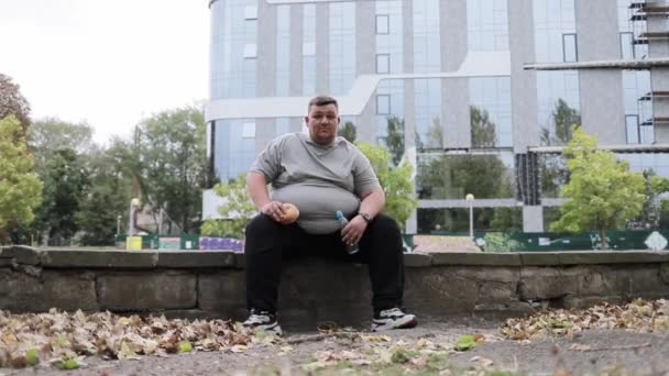 Retrato de un hombre con sobrepeso sentado afuera, comiendo un bollo y bebiendo agua. Un hombre gordo en el fondo de un edificio en la ciudad. - Metraje, vídeo