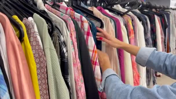 Una joven pobre navega por los bastidores de ropa en una tienda de segunda mano. Una joven elige ropa usada para ir de compras - Imágenes, Vídeo