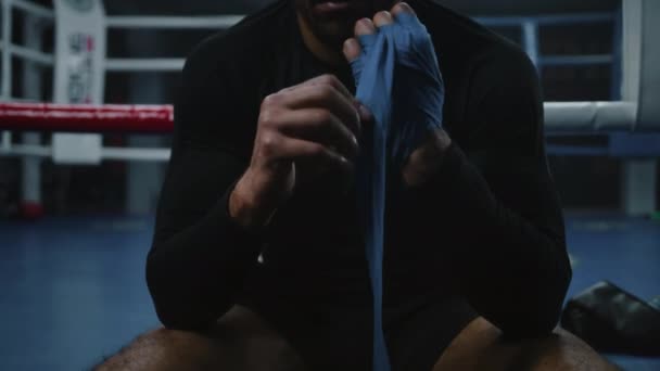 Афро-американский боксер сидит рядом с боксерским рингом и обмотывает руки бинтами для боевых искусств. Спортсмен тренируется в боксерском зале и готовится к бою или соревнованию. Физическая активность. Закрывай. Zoom out - Кадры, видео