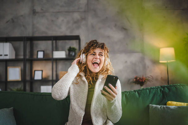 Eine erwachsene Frau aus dem Kaukasus mit Kopfhörern benutzt ihr Mobiltelefon zu Hause, um die Musik online zu hören, fröhliches Lächeln singt Freude haben Spaß allein Kopierplatz - Foto, Bild