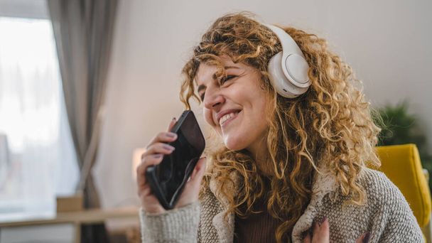 Kafkasyalı, kulaklıklı yetişkin bir kadın evde cep telefonu ile müzik dinliyor. Mutlu bir gülümseme. Keyfinize bakın. - Fotoğraf, Görsel
