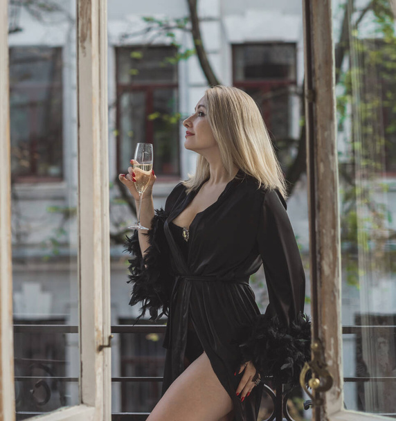 Femme élégante dans le boudoir. Femme européenne blonde en robe de soie noire avec des plumes sur une manche debout sur un balcon avec une coupe de champagne.   - Photo, image