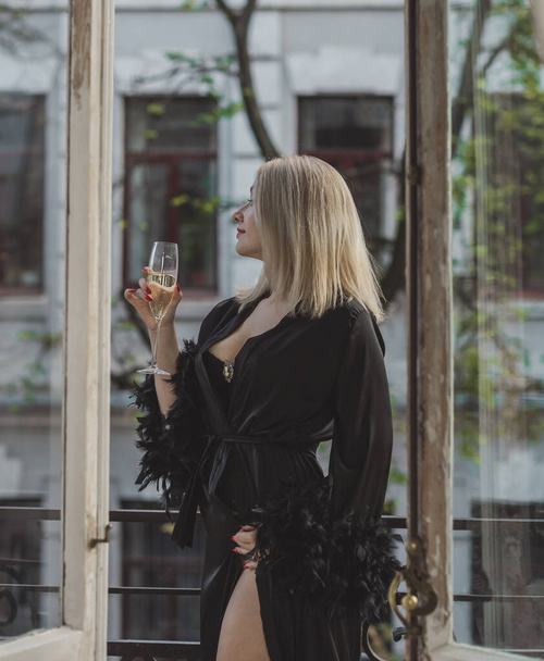 Elegante Frau im Boudoir. Blonde Europäerin in schwarzer Seidenrobe mit Federn am Ärmel, die auf einem Balkon mit einem Glas Champagner steht.   - Foto, Bild