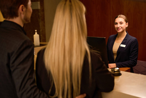 Χαρούμενη γυναίκα διαχειριστή στέκεται στο μετρητή υποδοχής και χαιρετισμό άνθρωπος και γυναίκα μετά την άφιξη στο ξενοδοχείο - Φωτογραφία, εικόνα