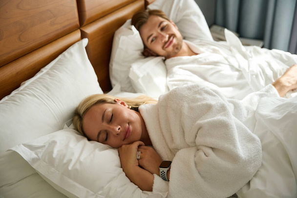 Giovane uomo si trova accanto a una bionda sonnecchiante, una coppia si riposa su un grande letto - Foto, immagini