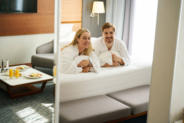 Os recém-casados felizes estão sentados em travesseiros em uma cama grande, café da manhã para dois é servido nas proximidades - Foto, Imagem