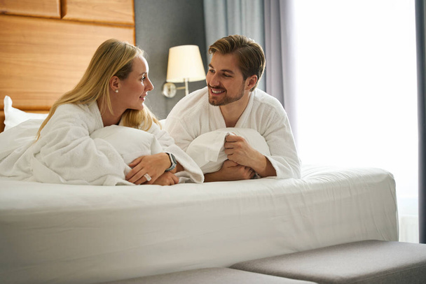 Frau und Mann kommunizieren auf einem großen Bett liegend, sie tragen Hotelfrottee-Bademäntel - Foto, Bild