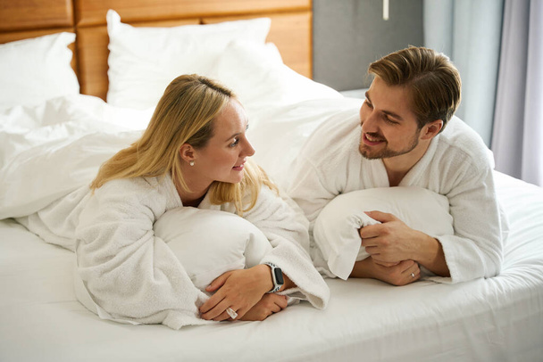 Guy ve kız arkadaşı büyük bir yatakta yatarken otel Terry cübbesi giyiyorlar. - Fotoğraf, Görsel