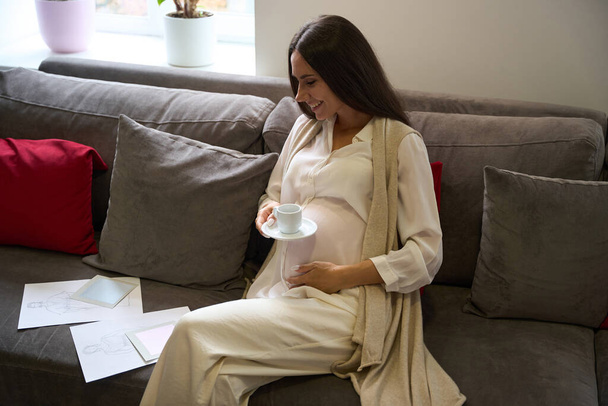 Encantadora mujer embarazada se sienta en una zona de relajación con una taza de café y bocetos de ropa - Foto, imagen