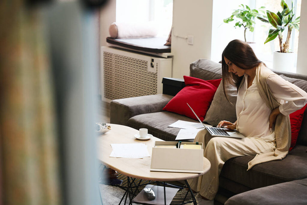 Designérka s bolestí zad pracuje na notebooku, sedí na velké pohovce - Fotografie, Obrázek