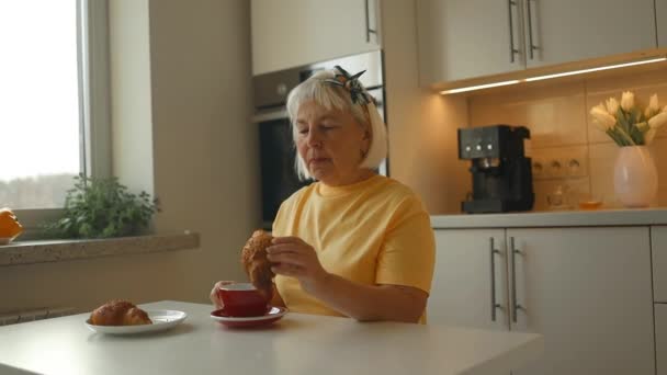 Aantrekkelijke energieke oudere blanke 60-jarige hongerige vrouw die croissants eet met chocolade als ontbijt op tafel in de keuken. Vrouwelijke drinken aromatische cappuccino koffie genieten ochtend ritueel - Video