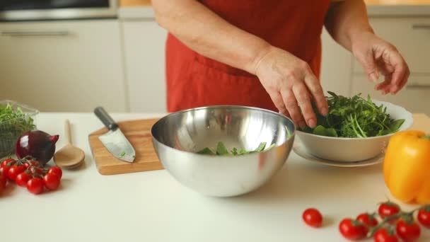 Lähikuva vanhempi nainen valmistelee kasvissalaattia keittiössä.Nainen lisäämällä arugula salaattia. Terveellistä ruokaa. Vegaanisalaattia. Kevyt. Laihdutussuunnitelma. Terveellistä elämäntapaa. Kotona kokkaaminen. Valmista ruoka - Materiaali, video
