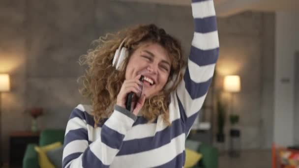 Una mujer caucásica mujer adulta con auriculares usar el teléfono móvil en casa para escuchar la música en línea sonrisa feliz cantar alegría divertirse solo copiar espacio - Imágenes, Vídeo