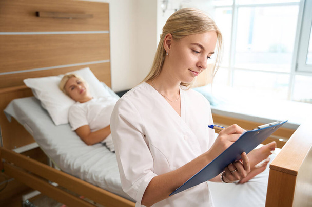 産科病院の医療ベッドに横たわるぼやけた妊婦の背景を持つクリップボードに焦点を当てた白人女性看護師のフロントビュー - 写真・画像