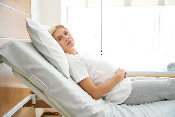 Vista lateral de una mujer embarazada europea adulta grave acostada en la cama médica, abrazando su vientre y mirando a la cámara en el hospital de maternidad - Foto, Imagen