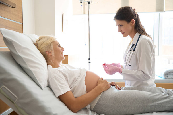 Ευρωπαϊκή γυναίκα νοσοκόμα προετοιμασία σύριγγα για ένεση και χαμογελώντας έγκυος καυκάσια γυναίκα στο ιατρικό κρεβάτι κοιτάζοντας ο ένας τον άλλο στην κλινική μητρότητας - Φωτογραφία, εικόνα